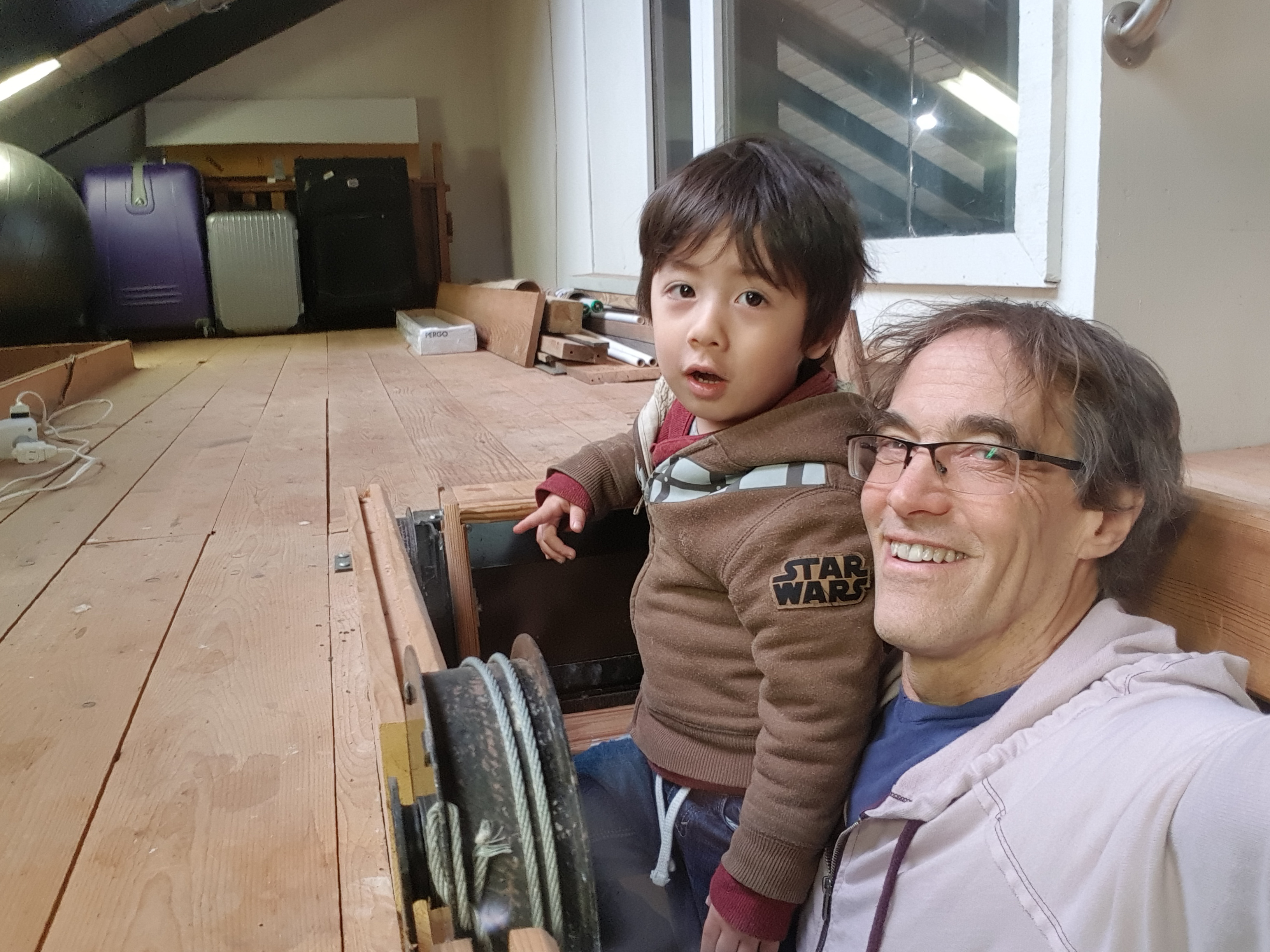 Smiling man with glasses and a young boy in a brown jacket beside a wooden deck; a wheel-and-gear device sits in the foreground.