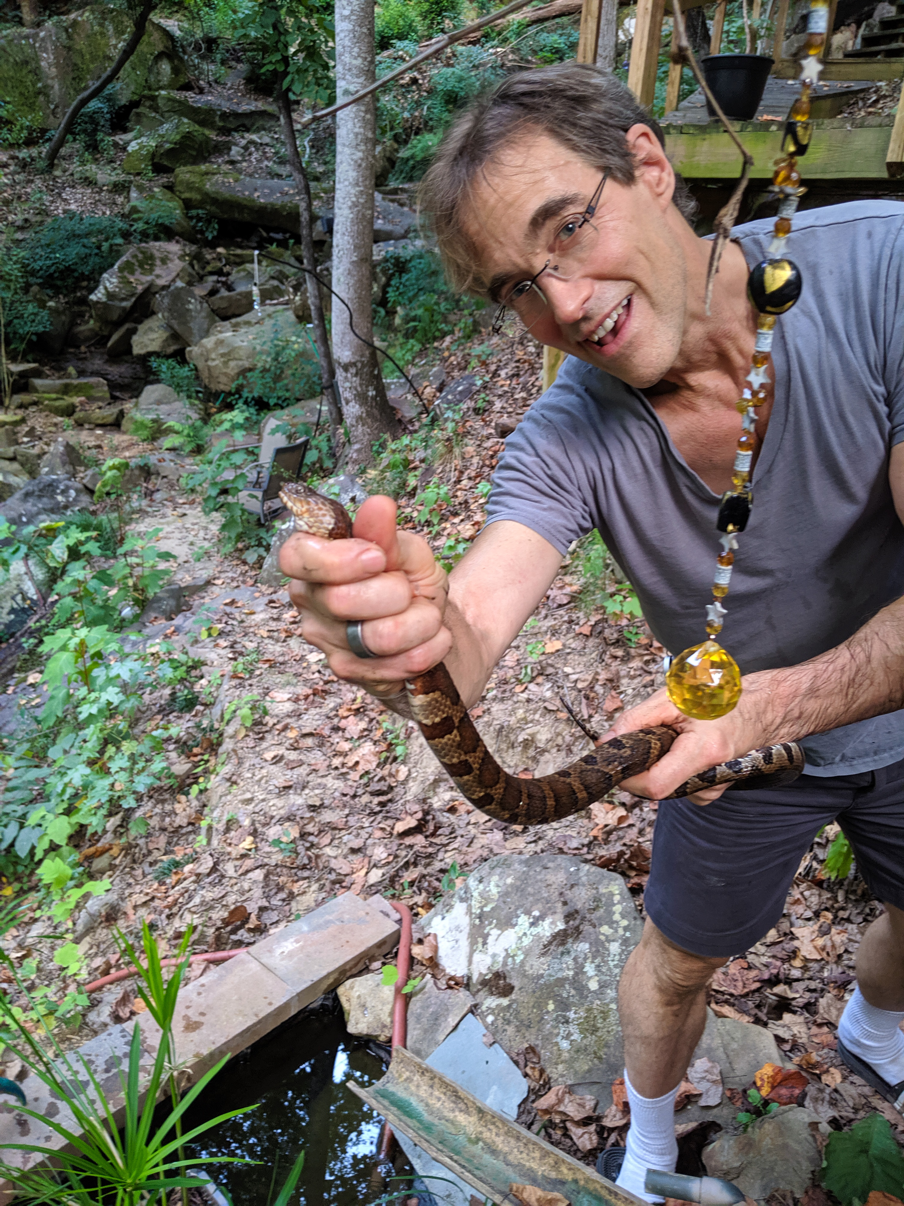 Smiling man in a gray shirt holds a brown patterned snake over a small stream in a leafy setting, wearing a necklace with a yellow pendant.