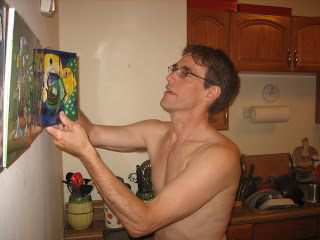 Shirtless man with glasses reaches toward a bright, abstract painting on a kitchen wall; warm wood cabinets and cluttered counter behind.