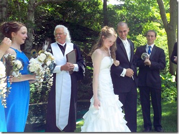 Bride in a white wedding dress walks outdoors; officiant in a black robe beside her as bridesmaid in blue holds a bouquet and guests in suits watch.