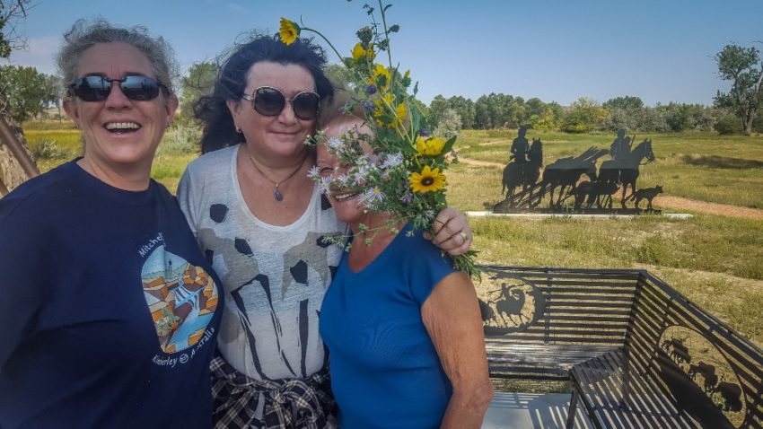 Three women pose on a sunny deck; one holds a bouquet of yellow flowers, with black poodles in a fenced field behind them.