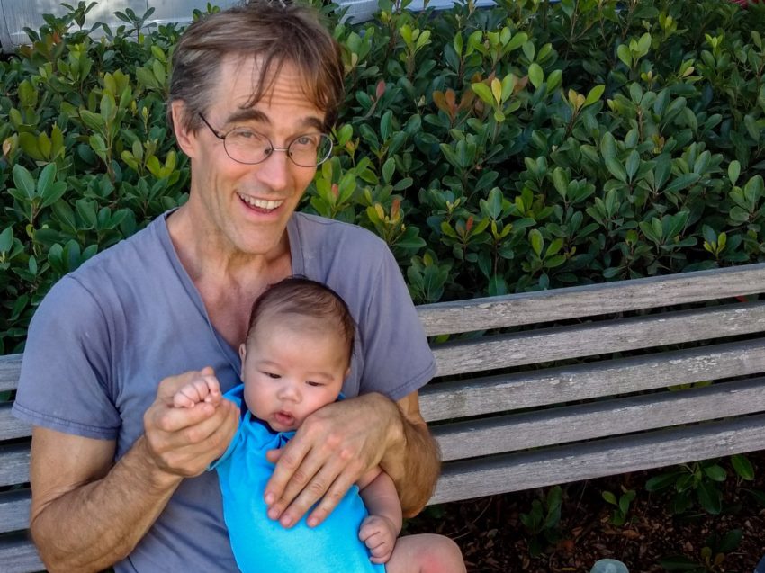 Man with glasses in a blue shirt sits on a wooden bench, cradling a baby in blue, with green bushes behind.