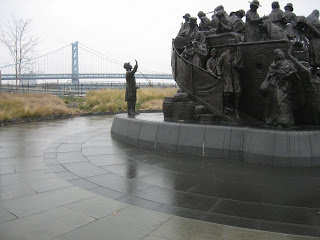 Bronze soldiers statue on a circular pedestal; a lone man in light clothing stands nearby as a bridge spans a river under a gray sky.