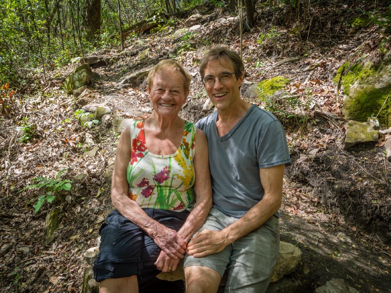 Two smiling adults sit close on a mossy rock in a sunlit forest; the woman wears a colorful floral top, the man a gray tee.