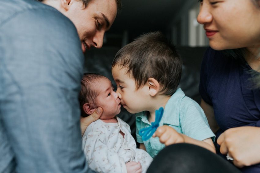 Dad leans toward a newborn as a toddler in teal noses the baby; a smiling woman sits nearby, watching.