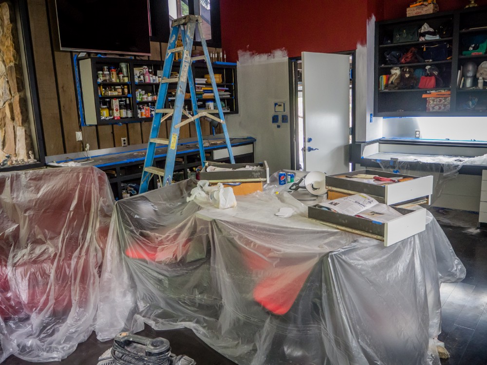 Construction workshop with plastic-covered tables, blue ladder in center, wall shelves of cans, open white door, red wall accents, scattered tools.