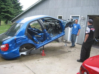 Blue compact car with doors removed and interior exposed, raised on red jacks beside a gray metal workshop; two men watch.