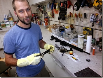 Man in blue T-shirt and yellow gloves at a white workbench in a busy workshop, holding a tool over a long black object.
