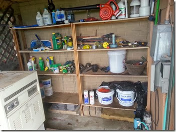 Wooden shelf filled with cleaning sprays, paint cans, buckets, and tools; white laundry appliance to the left; orange hose reel on top.