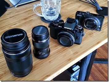 Two black cameras with lenses on a wooden table; a large Olympus lens on the left, a mid-size lens beside it, a glass mug behind, blue cables along the edge.
