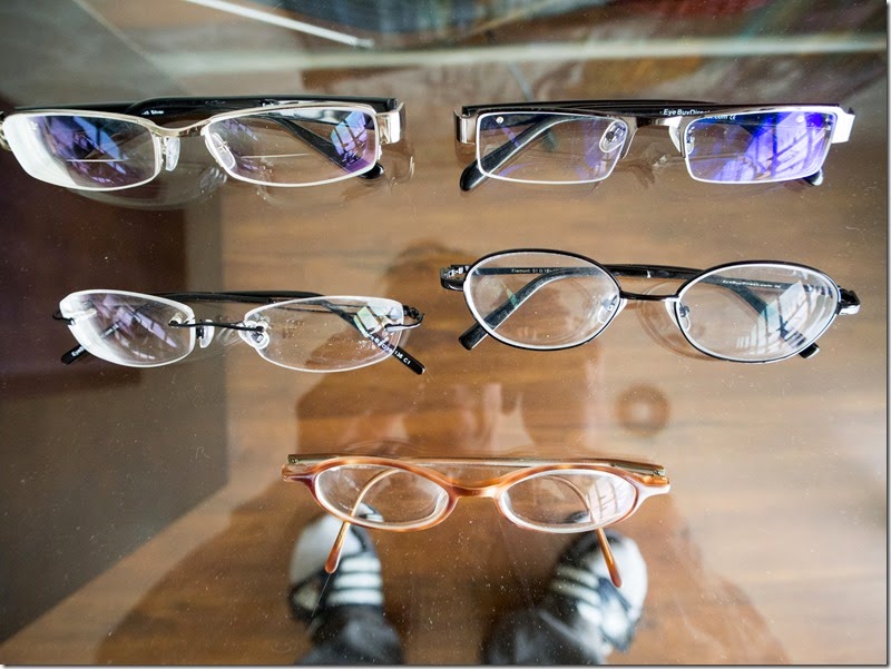Multiple eyeglasses arranged on a wooden display, with metal and plastic frames in white, silver, black and gold, reflected in a glass surface.