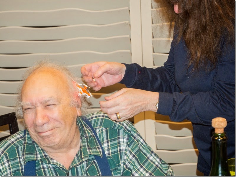 Elderly man in green plaid shirt smiles as a person in blue hands him a small object; a bottle rests on the table, blinds behind.