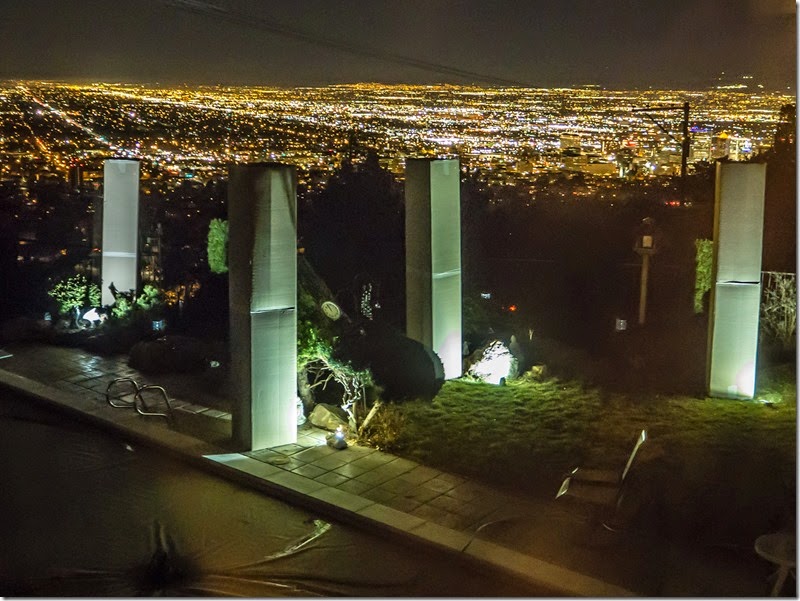 Night view of tall white concrete columns forming a box-like frame atop a hill, with golden city lights in the distance and a landscaped foreground.