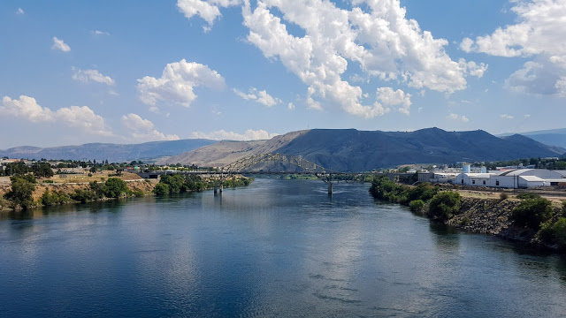 Wide river with calm blue water, green banks, distant hills, and a bridge crossing the water under a partly cloudy sky.