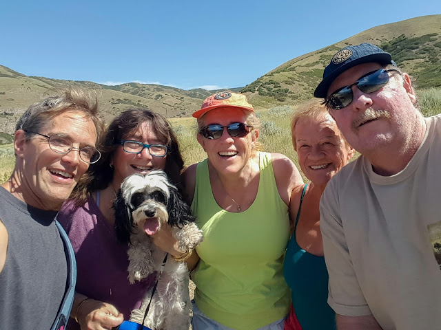 Smiling group of five adults and a small fluffy dog taking a selfie outdoors, with green hills and blue sky behind them.