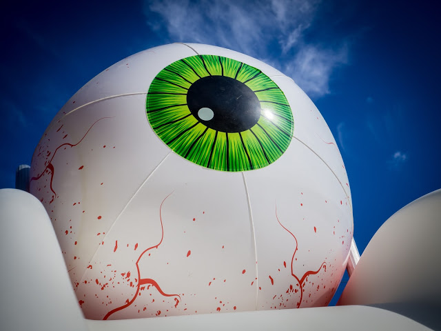 Giant white eyeball sculpture with a bright green iris and red veins, set against a blue sky and surrounded by white inflatable shapes.
