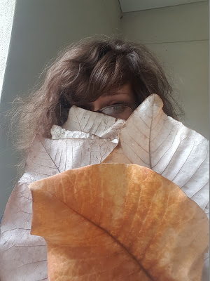 Person with brown hair peeks over a large orange seashell, wearing pale clothing, against a light wall.