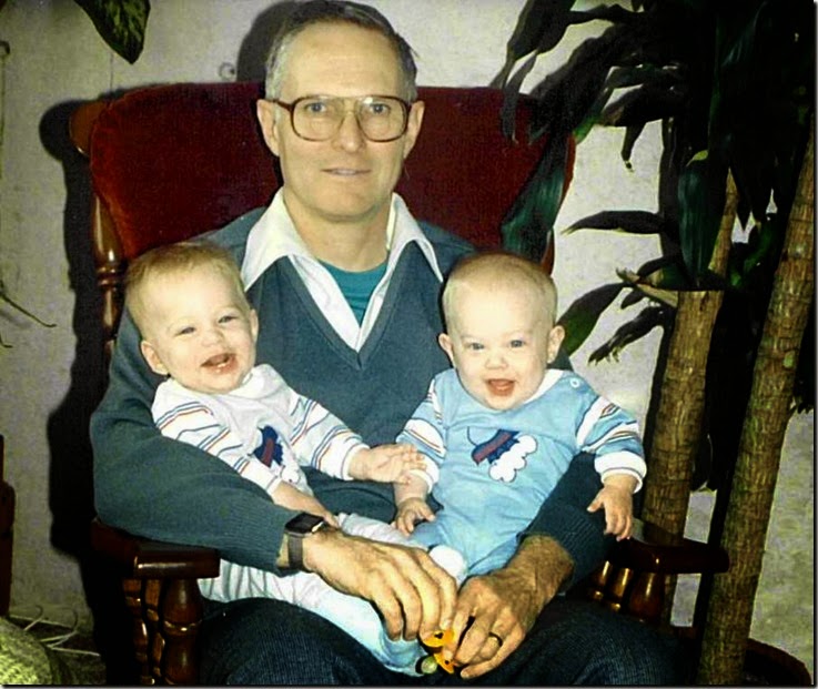 Older man with glasses sits in a dark wooden chair, cradling two smiling babies in blue outfits.
