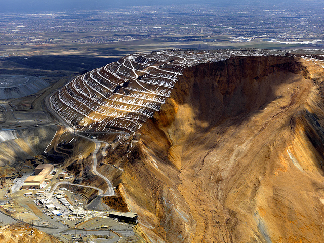 Terraced open-pit mine in orange-brown rock with winding roads along the benches under a blue sky.