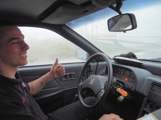 Young man driving a gray car, thumbs-up with his left hand, dashboard and steering wheel visible, road ahead through the windshield
