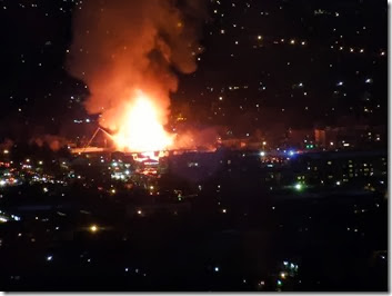 Nighttime cityscape with a large orange fire and smoke plume rising above buildings, orange glow illuminating surrounding lights.