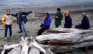 Six people in colorful jackets on a cloudy shoreline with large driftwood logs, posing with outstretched arms.