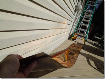 Beige horizontal siding with a green ladder leaning against the wall, sunlit wooden deck, and a hand holding a reflective metal sheet.