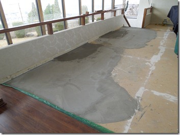 Sunlit sunroom with a beige floor, large gray concrete patches, wooden trim, windows along the left, and a doorway at the far end.