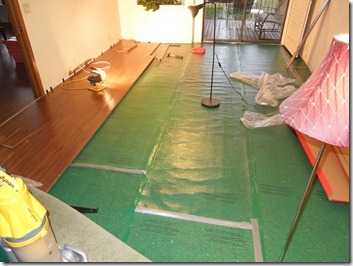 Long hallway with green floor covering over a wooden floor, central metal pole, pink draped cloth on right, bags and tools along edges.