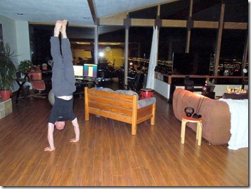Person in dark clothing performs a handstand against a vertical pole in a living room with wooden floor, brown sofa, and blue ceiling.