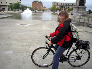 Smiling woman in a red-and-black jacket sits on a black bicycle with a backpack in a wide gray plaza, a fountain to the left and buildings behind.