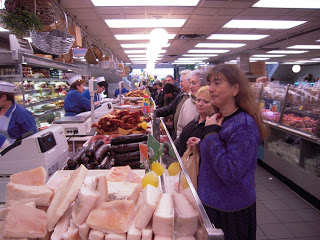 Supermarket deli with cheese blocks in foreground; woman in purple with a child at center, shoppers and meat counters along both aisles.