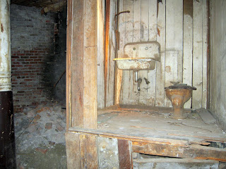 Rusty sink mounted on a weathered wooden shelf with a curved faucet; worn planks fill the frame, a brick wall and a shadowy doorway visible to the left.
