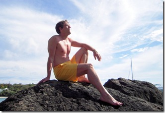 Shirtless man in yellow shorts sits on dark volcanic rocks by the sea, gazing upward, with a bright blue sky and wispy clouds behind.