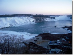 Niagara Falls area: wide river with white cascades along the cliff, a viewing platform and buildings at the water’s edge under a dusky blue sky.