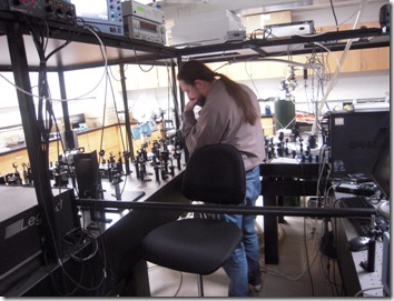 Man in a gray jacket stands at a cluttered control booth above a crowded arena, with tangled cables, a black chair, and equipment visible.