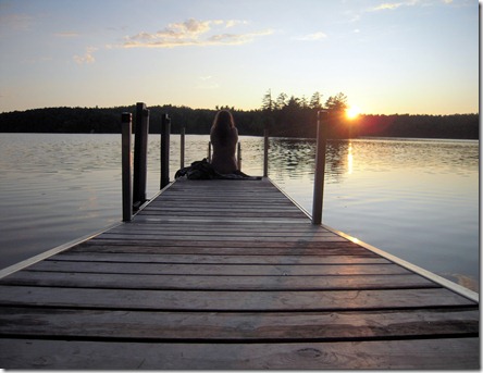 Wooden dock extends into a calm lake at sunset; a person sits at the far end, orange light reflecting on the water, trees in the distance.