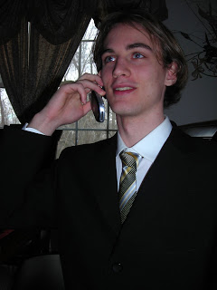 Young man in a dark suit with a striped tie, holding a cell phone to his ear indoors; dark curtains and a window behind him.
