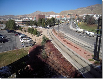 Blue sky over a campus with curved tram tracks beside a road, parked cars on the left, and distant modern buildings with brown hills beyond.
