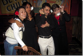 Five friends posing together in a dim venue; center man in black shirt and white pants, flanked by others in dark outfits.