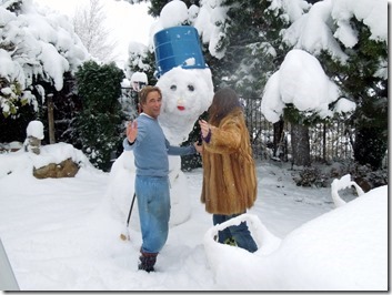 Two people pose with a large snowman wearing a blue top hat in a snowy yard; one in blue, one in brown coat, snow-covered trees behind.