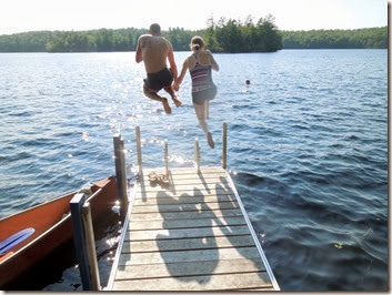 Two people jump from a wooden dock into a blue lake; a red boat rests to the left, with green trees and a sunny sky in the background.