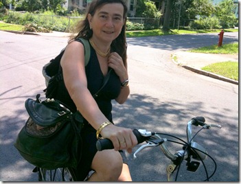 Woman with dark hair in a sleeveless black top sits on a bicycle along a sunny suburban street, a black shoulder bag over her back.