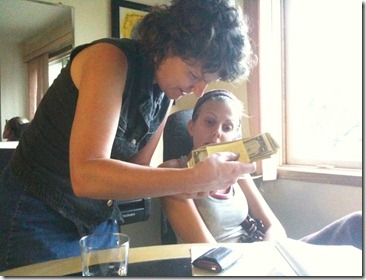 Standing woman with short dark hair in a black sleeveless top leans over a table, examining a yellow card; another woman sits by a sunlit window.