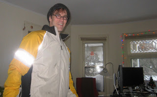 Man in white and yellow jacket stands in a room with a desk, computer monitors, and festive string lights by a window.
