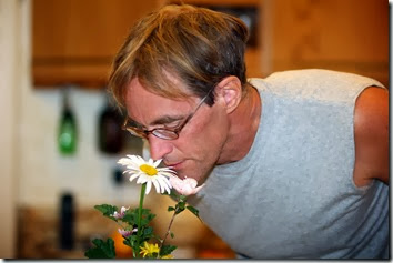 Man in a gray sleeveless shirt leans toward a bouquet of white daisies on a kitchen counter, with warm wood cabinets in the background.