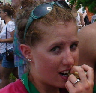 Close-up of a woman with teal sunglasses on her head and pink top, biting a snack amid a crowded marathon.