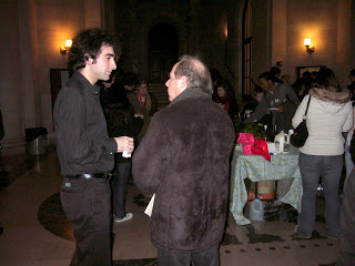 Two men chat at a warm-lit indoor event; one in a black shirt, the other in a brown jacket, with guests and tables in the background.
