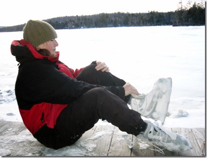 Person in a red jacket and green beanie sits on a snowy dock by a frozen lake, with a white ice skate near their feet.