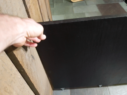 Hand grips a dark rectangular panel attached to a wooden frame, with a tiled floor in the background.